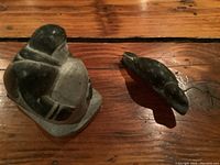 Two dark soapstone carvings on a wooden surface: a seated human figure and a bird, both with worn surfaces.