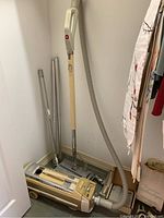 View of gold and cream Electrolux vacuum cleaner upright with hose and wood-handled extension wand leaning, several chrome metal extension tubes beside it