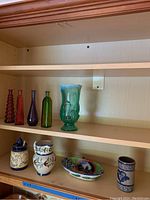 Five colored glass vases on upper shelf and four pottery pieces on lower shelf inside wooden cabinet.