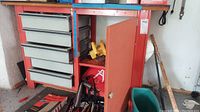 Front view of red framed workbench cabinet with multiple grey drawers and a storage door open showing internal items.
