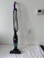 Full front view of the vacuum cleaner showing its upright structure, purple handle, power cord wrapped around the rear, and translucent green dust compartment.