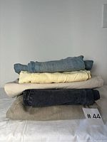 Stack of five folded pants including denim jeans, yellow linen pants, beige and grey pants.