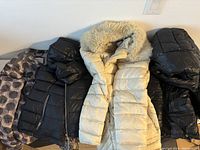 Four coats laid out including cream puffer with fur hood and three black coats, one with patterned lining.