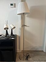 Floor lamp with brushed metal base and white pleated lampshade standing against a white door.