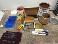 Wide view of jewelry making supplies including container of red sand, wooden blocks, glue tube, metal rods, black metal plates, and blue sticks.