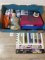 Top view of box showing assorted office supplies including acrylic paint bottles, rulers, pencils, correction tape, binders, and packs of pens and markers.