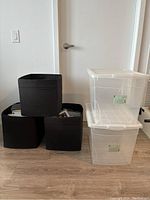 Three black fabric storage bins stacked in a pyramid shape next to two clear plastic storage containers with snap-on lids placed vertically on the floor near a white door.
