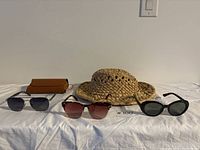 Three pairs of sunglasses aligned on a white surface along with a brown leather case and woven straw hat.