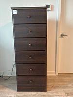 Front view of tall dresser showing six drawers with round knobs, dark wood finish, standing on floor beside a wall.