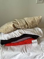 Stack of five women's garments folded, showing a beige textured fabric item on top, followed by white, black, red and white patterned, and cream fabric items.