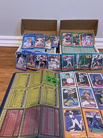 Two cardboard boxes filled with various baseball cards arranged in sleeves and stacks on the floor against wall.