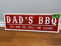 Front view of a rectangular metal sign with 'DAD'S BBQ' and 'THE MAN, THE GRILL, THE LEGEND' in red on a white background.