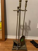 Full view of the fireplace tool set including stand with four tools propped up across it on wooden floor