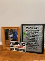 Three wall art pieces arranged on the floor: framed graphic plaque, metal 'Man Cave' sign in black frame, and two wooden block signs with motivational quotes.