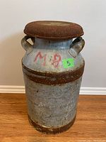 Photo front view of rusty metal milk can showing rust on lid and metal body with red markings