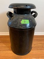 Black metal milk can with lid and two handles shown frontally.