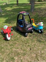 Three ride-on toys displayed on grass: black Batman car in center, red motorcycle on left, blue scooter on right.