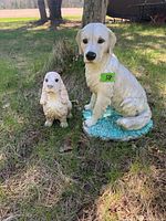 Front view of cream-colored dog statue and smaller white bird statue sitting together on a green base outdoors.