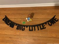 Photo showing the black arched sign with text 'DREAM BIG LITTLE ONE' laid flat on a wooden floor next to the beaver bank figurine.