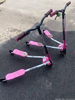 Front angle view of two pink and white Sporter wiggle scooters on asphalt ground showing the foot platforms and handlebars.