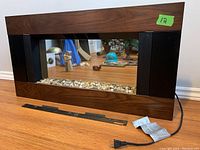 Front view of the electric fireplace showing wood grain frame, black side panels, clear front window with decorative river stones inside, a power cord, and a metal mounting bracket below it
