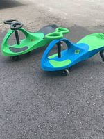 Two plastic Wiggle Bikes on asphalt; one green and black, one blue and green, showing side and front views of the ride-on toys.