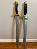 Two wooden swords standing upright against a white wall; one with gold-painted pommel and guard, one with silver.