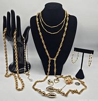 Full display of multiple gold tone necklaces, earrings, and stick pin arranged on black jewelry stands and hands. Shows variety of chain styles and pendant designs including heart earrings, red stone stick pin, and pearl necklace.