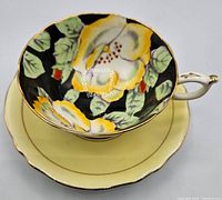 Wide mouth cup and saucer showing detailed hand-painted yellow and white dogwood flowers with jewelled petals and green leaves on black background, with gold rims.