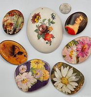 Front view of seven mixed vintage brooches arranged with a quarter for size reference, showing colorful floral designs and a portrait brooch.