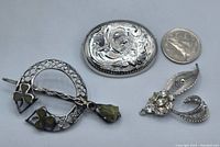 Three silver brooches next to a quarter for size reference: circular shamrock pin, oval floral Birks brooch, and rhinestone AmyLee brooch