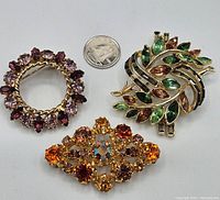 Top-down view of the three vintage rhinestone brooches with a quarter for scale, showing one green and orange leaf motif brooch, one round purple brooch, and one orange decorative brooch.
