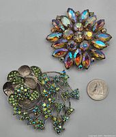 Two vintage rhinestone brooches side by side with a quarter for scale, one green floral and one round multi-colored