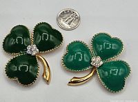 Front view of two three-leaf clover brooches with green glass leaves and rhinestone centers. Quarter placed nearby for size reference.