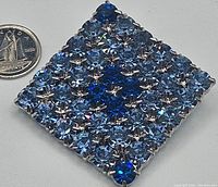 Front view of a square vintage brooch featuring blue rhinestones in multiple shades arranged in a grid, placed next to a coin for size reference.