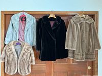 Four vintage faux fur coats and jackets hanging on brown wooden doors showing front views with collars, varying lengths and colors: light blue, tan brown, black, beige.
