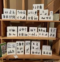 Overall shelf display of approximately 26 white Dept. 56 boxes with grayscale illustrations of figurines