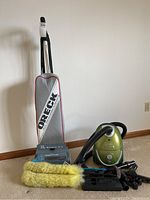 Oreck upright vacuum and Royal canister vacuum with multiple vacuum attachments including yellow mop head, brush, nozzles, hoses, and extension tubes on floor against wall