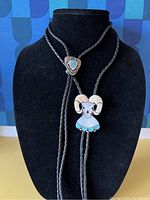 Full view of two bolo ties on black display background; a ram skull design with turquoise stones and a shield-shaped turquoise stone with textured silver.