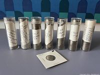 Seven plastic coin tubes containing US circulated nickels stacked inside with handwritten paper labels indicating date ranges. One coin in a white cardboard holder is positioned in front.