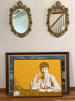 Two oval gold-tone plastic mirrors mounted on wall above two framed paintings (Bill Berry portrait and waterfall forest scene)