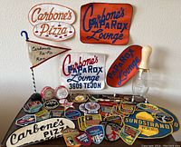 Photo showing vintage embroidered patches and promotional towels for Carbonne's Pizza and Caparox Lounge along with assorted pins and keychains on a dark table.