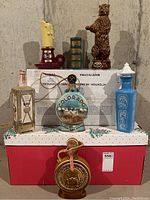 Full view of seven vintage decanters arranged with red and white holiday-themed box under the group, some standing on a cardboard box in the background. Varied designs with ceramic and glass materials evident.