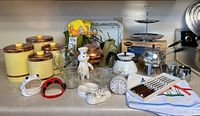 Wide view of kitchen countertop showing all vintage kitchenware items including the canisters, fondue pot, pastry press box, measuring cups, ceramic accessories, fork set, glassware, kitchen towels, and serving tray.