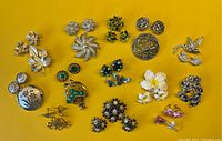 Overhead photo showing multiple vintage pins and matching earring sets laid out on a yellow textured surface, various metal tones and colors visible.