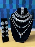 Frontal view of four rhinestone necklaces displayed on a black velvet bust and four pairs of clip-on earrings on a stand to the left.