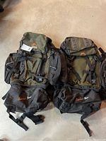 Two green and black Outdoor Gear backpacks side by side showing front with multiple compartments and visible logos.