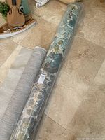 Two rolled woven carpets on a stone floor, one floral grey rolled wrapped in plastic, the other textured grey rolled carpet partly unrolled.