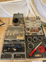 Photo showing all items included in the lot: Ryobi rotary tool in case, assortment of sanding drums, grinding stones, saw hole blades, diamond cutter, and other accessories laid out in multiple plastic cases on wooden table.
