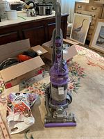 Full view of the Dyson DC15 Animal upright vacuum in purple and gray on a carpeted floor, showing the main body and attached hose, with background room items.